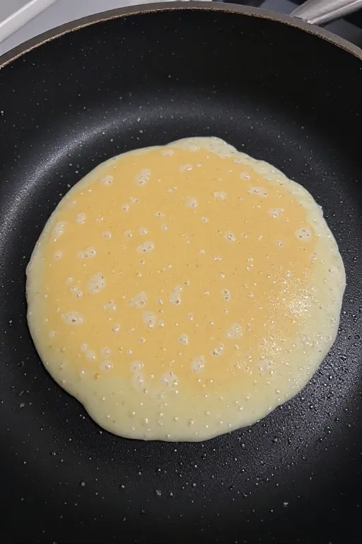 Fluffy pancake cooking in pan with bubbles forming on surface ready to flip